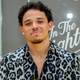 Anthony Ramos, dressed in a silver and black faux leopard print suit, smiles in front of an In the Heights promo board.